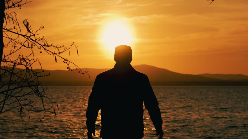 The silhouette of a single man standing on the shore of a lake at sunset. A happy person enjoys life, achievements and success by raising his hands in the air. Rear view.	