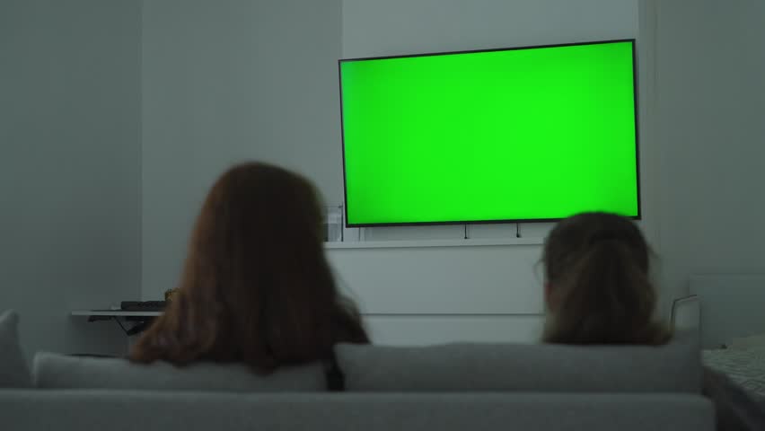 Two people sit on a sofa in a modern living room watching a large tv with a bright green screen, seen from behind in a calm home setting.