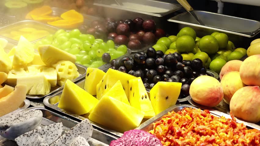 A lively Chinese night market fruit stand with steaming trays of freshly cut tropical and seasonal fruits like watermelon, dragon fruit, kiwi, and strawberries under warm lights.
