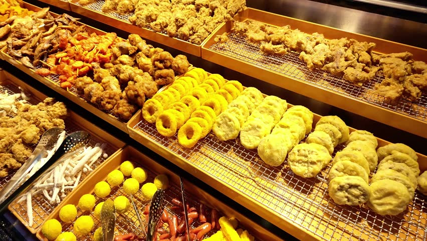 Close-up panning shot across a well-lit Chinese night market food display featuring crispy fried chicken, squid, onion rings, buns, and assorted street snacks.