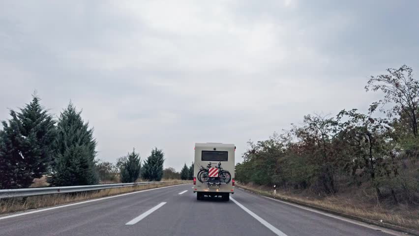 Camper van with bicycles mounted on the rear drives along a highway at dawn. The road is clear, flanked by trees and a guardrail under an overcast sky, depicting an authentic travel moment.