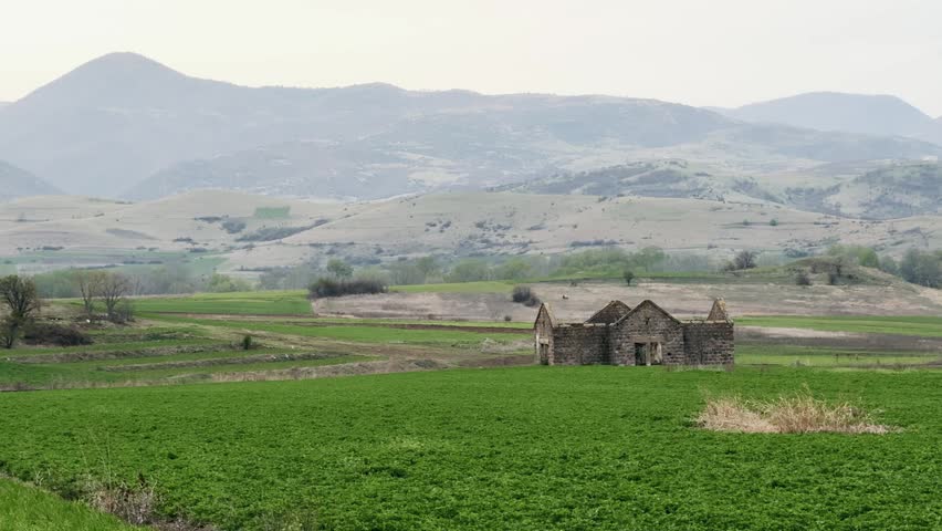 A dilapidated stone building stands prominently in a vibrant green field, flanked by rolling hills and distant mountains under a cloudy sky.
