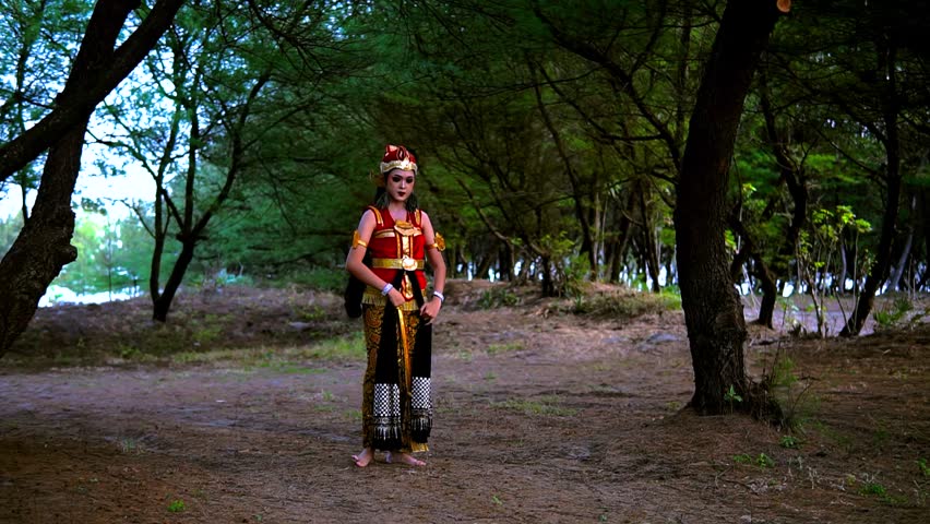 Woman in ornate traditional costume performs graceful dance sequence along dirt path inside shaded forest park, moving from still pose into flowing gestures