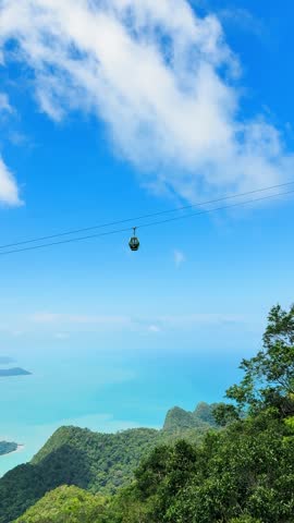 4K Vertical shot of cable car above green forest and in front of turquoise blue colored sea and sky with clouds at Langkawi, Malaysia. Scenic travel background. Summer vacation concept.