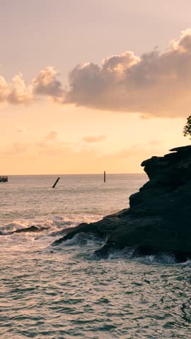 4K Vertical shot of ocean waves hitting rock during sunset at Langkawi Island, Malaysia. Orange clouds in sky during sunset. Sun setting above the Strait of Malacca with little islands of Langkawi. Nature background.
