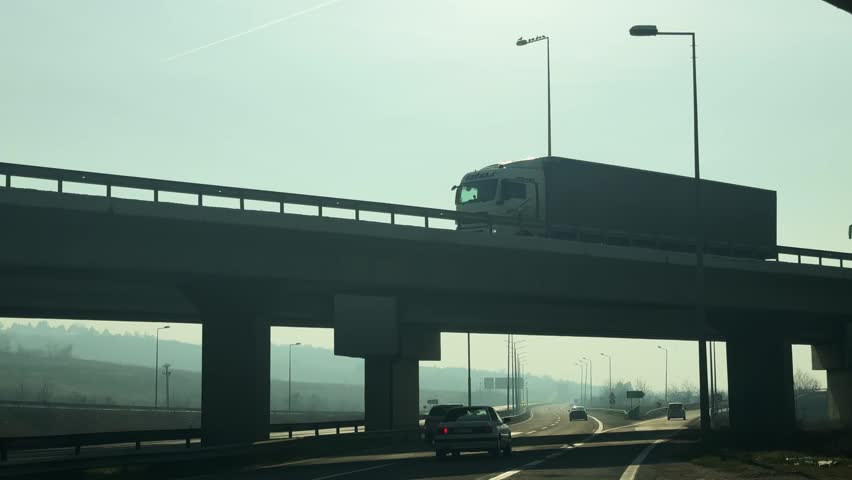 View of a multi-level highway with various vehicles including a bus, truck, and cars traveling on both the elevated overpass and the road below, under a hazy sky.