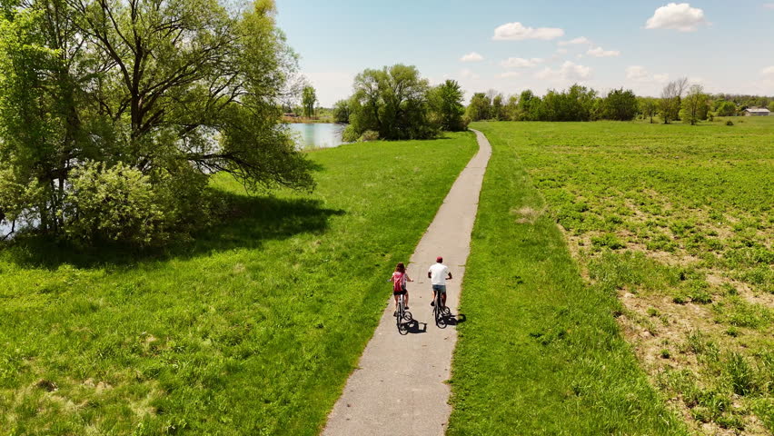 Couple cycles paved path through nature with water views, St Lawrence Parks, Ontario. Active lifestyle, healthy living, outdoor recreation, Canadian landscape.