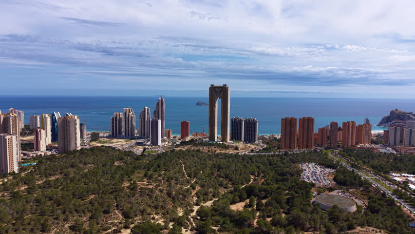 Panoramic view of Benidorm, Spain, showcasing iconic skyscrapers including the Intempo tower, lush hillside vegetation, and the blue Mediterranean Sea under cloudy sky