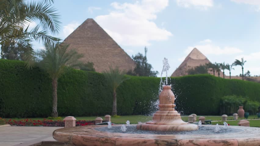 Slow motion 4K travel video featuring a fountain in the foreground with the Pyramids of Giza in the background, surrounded by palm trees and lush gardens in Egypt.