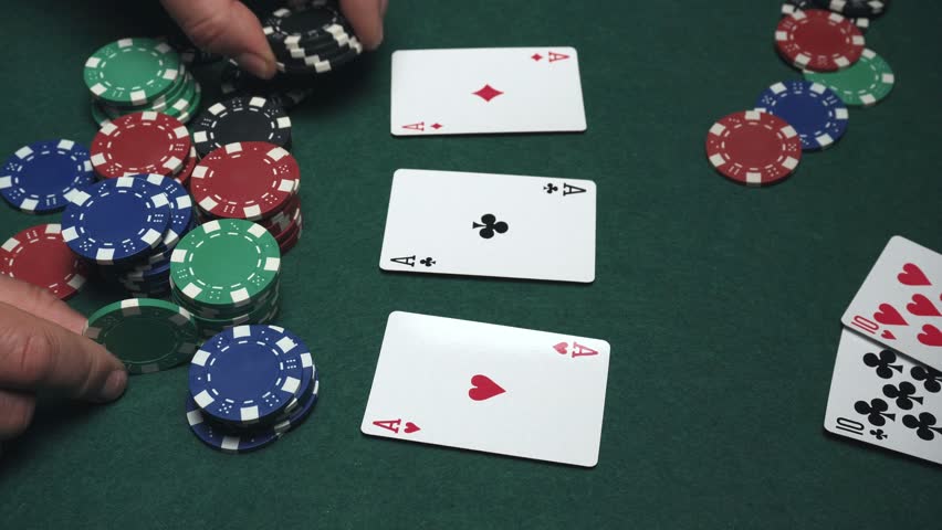 Hands carefully stack black poker chips on top of playing cards displaying aces, with a variety of colorful chips scattered across a green felt table in a casino environment