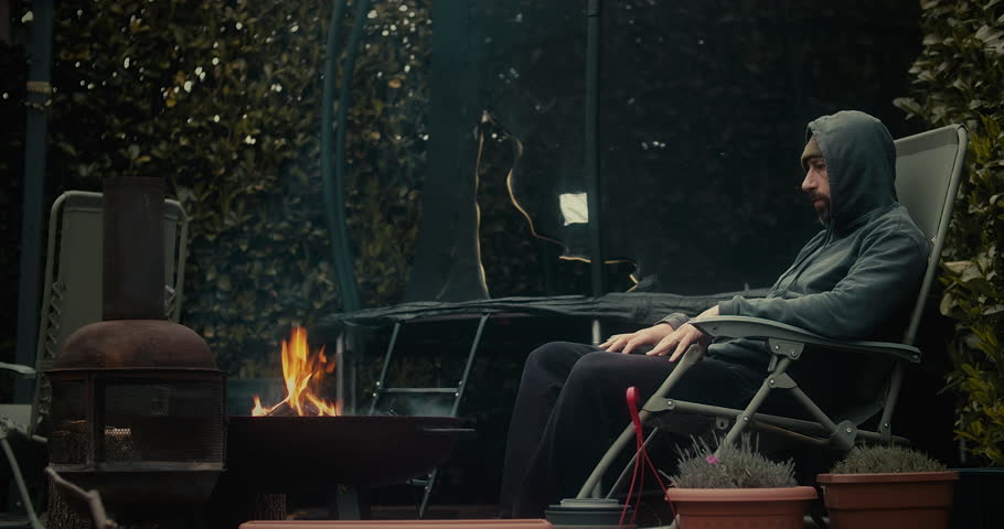 Man sitting in front of a fire pit, alone outside, starring at a yellow orange flame.