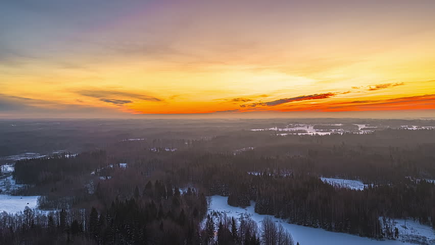 Aerial sideways drone hyperlapse glides over vast snow-covered boreal forest with frozen lakes, ground fog and vibrant orange-red sunset sky