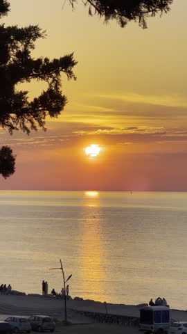 Golden sunset over the Mediterranean Sea at Hadjret Ennous, Tipaza, Algeria. Calm water reflects the warm orange sunlight while people enjoy the coastal scenery during a peaceful evening.