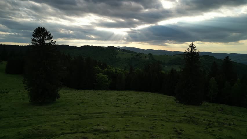 Captivating panoramic view of a lush green landscape featuring rolling hills and dense forests under a dramatic, cloudy sky with striking sun rays breaking through, creating a serene and ethereal atmosphere. Paltinis, Sibiu, Romania