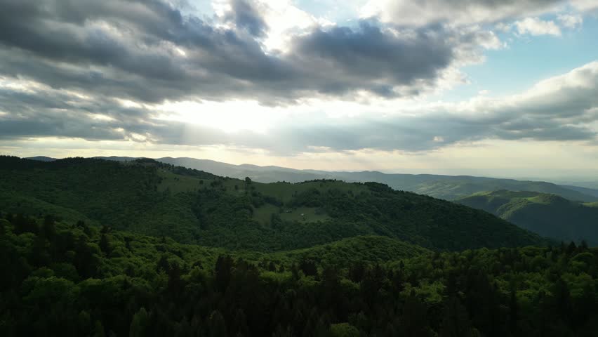 Expansive panoramic view of rolling green hills and lush forests under a dramatic sky with sun rays piercing through dark clouds. A peaceful natural landscape evoking tranquility and untouched wilderness. Paltinis, Sibiu, Romania