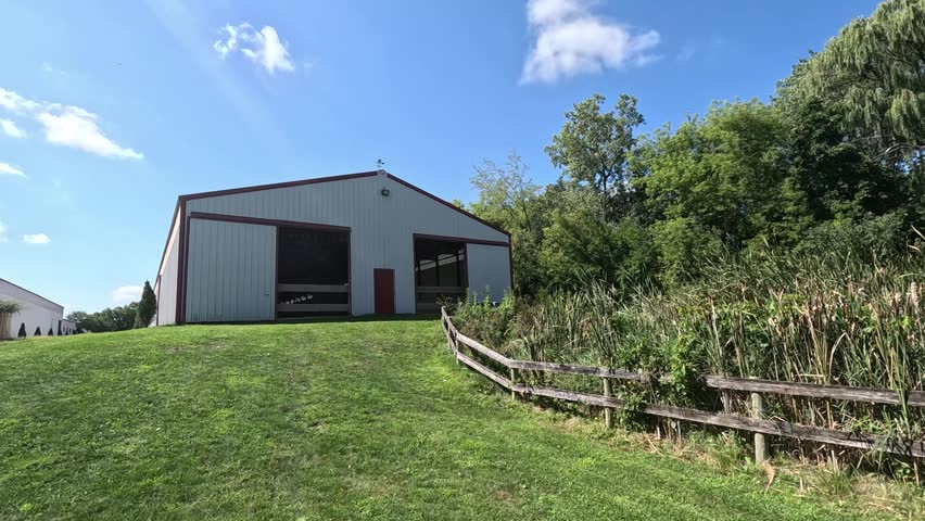 A barn on a summer day surrounded by open sky, trees, wooden fences, and plants.
