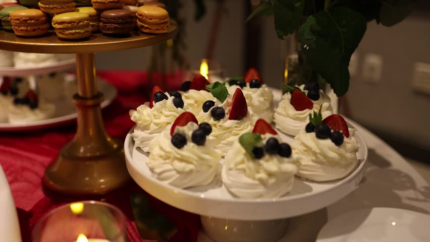 Plate of whipped cream pavlovas topped with fresh strawberries, blueberries and mint, beside colorful macarons on a stand at a warm indoor dessert table.