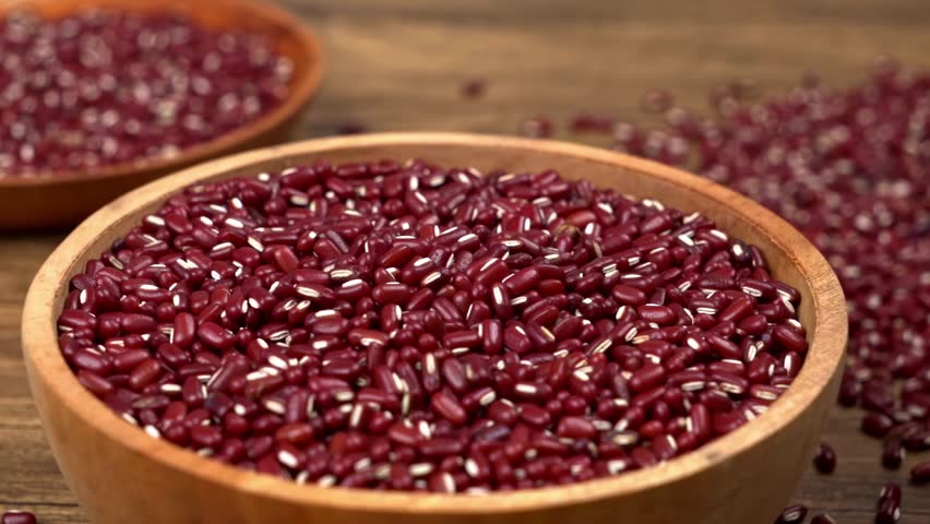 A wooden spoon is used to scoop and pour vibrant red adzuki beans into a rustic wooden bowl. The scene highlights the rich color and texture of the healthy legumes on a natural wooden surface.