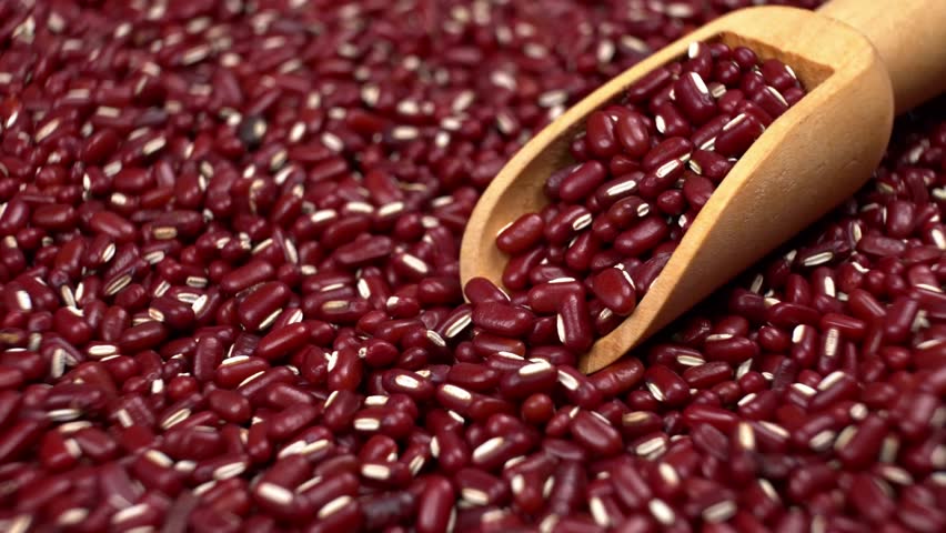 A close-up shot captures vibrant red adzuki beans being poured from a wooden scoop. The beans fall and scatter, creating a dynamic visual of food preparation.