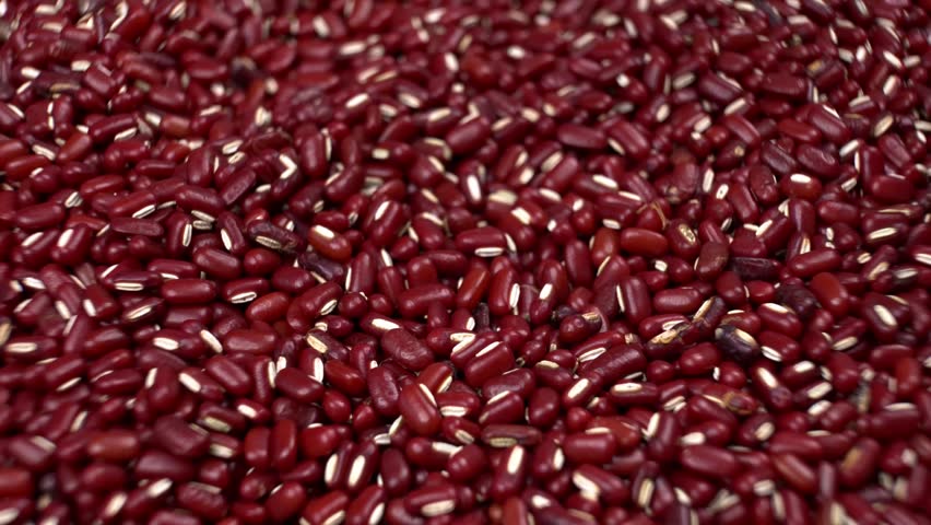 A wooden spoon scoops and pours vibrant red adzuki beans, highlighting their uniform color and smooth texture in a close-up, full-frame shot, emphasizing freshness and quality for culinary use.