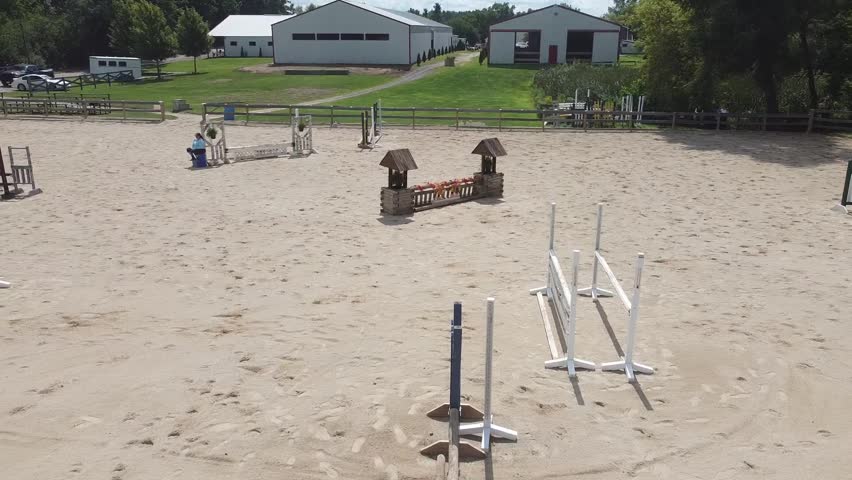 A drone shot of an outdoor arena and barn as an equestrian and her horse leap over jumps.