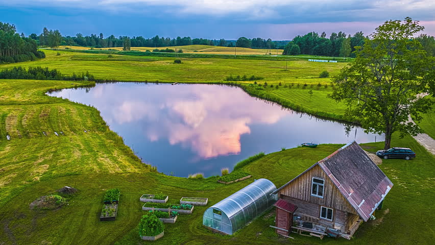 Timelapse of a quiet rural homestead beside a reflective pond at sunset, showing shifting light over fields, a greenhouse, a wooden house and calm countryside surroundings.