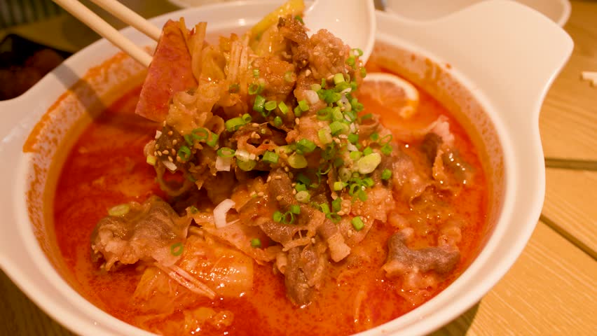 Close-up shot of chopsticks lifting noodles from a steaming bowl of spicy red beef broth.