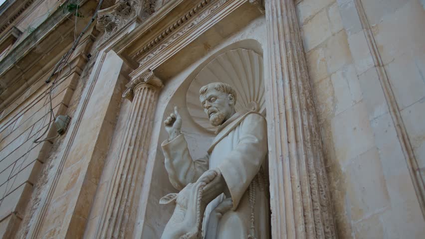Franciscan saint sculpture carved in Lecce stone decorates a Baroque church façade in Lecce, Lecce, Apulia, Italy, showcasing religious art and local architectural heritage, upward pan reveal shot.