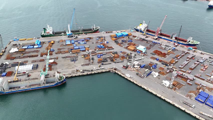 Drone footage of a cargo terminal in Busan Port, South Korea. Docked vessels, port cranes, steel materials, industrial storage areas and maritime logistics infrastructure along the waterfront.