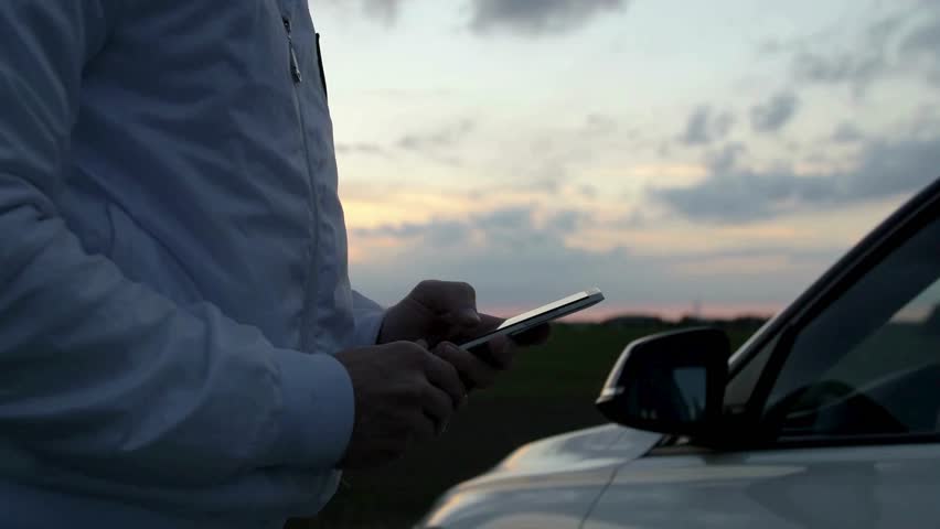 Person stands near a white car, using a smartphone during sunset. Sky is orange and cloudy. Focus on hands and phone with car in the shot.