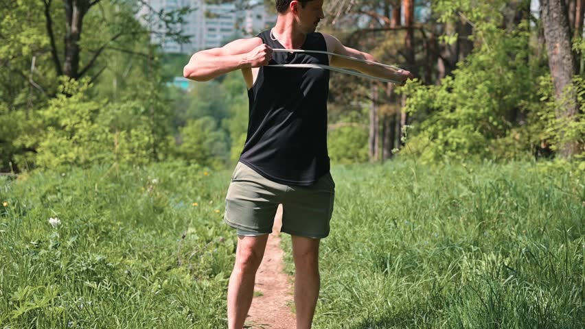 A person performs stretching exercises using a resistance band in a natural park setting with green grass and trees.