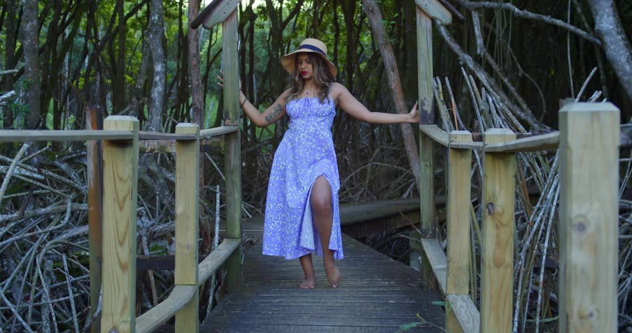 Indian girl laughing and spinning on a rustic boardwalk; her blue floral dress flares against a backdrop of towering pines.