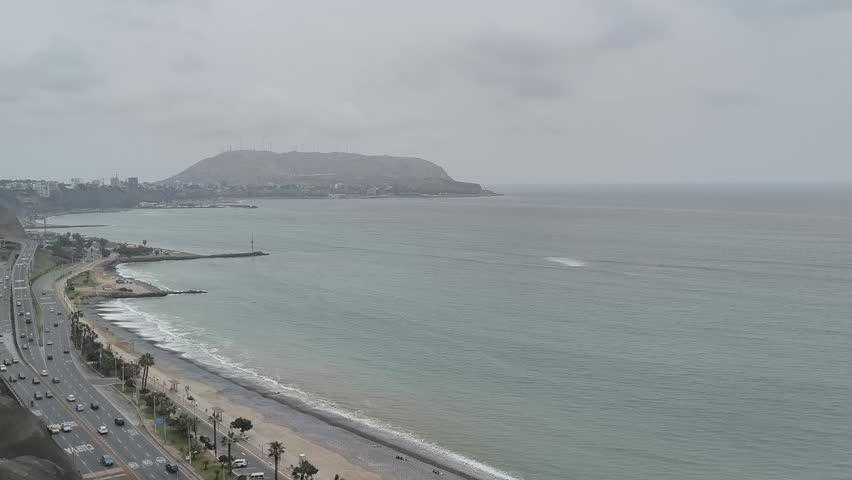 High-angle panoramic view of the coastline in Lima, Peru, featuring the Pacific Ocean, a coastal highway with moving traffic, and the iconic Morro Solar in the background under a cloudy sky