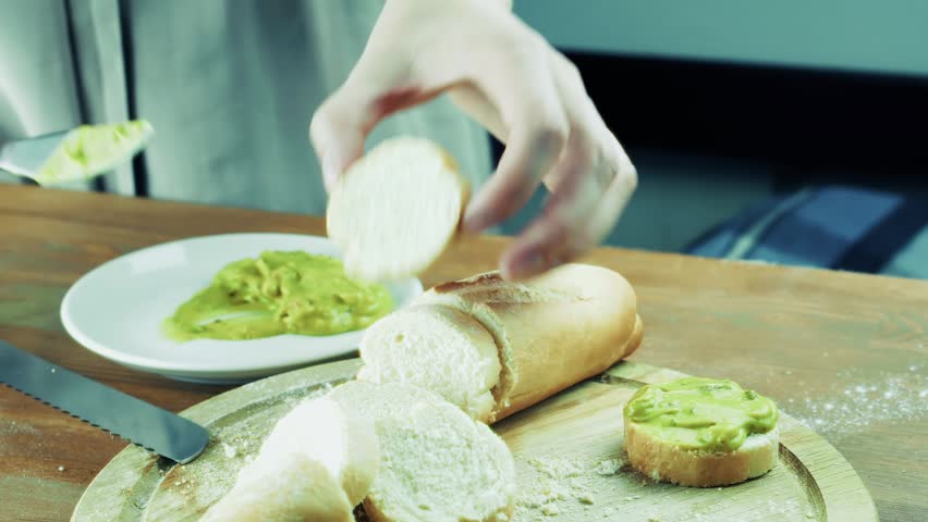 ands use a table knife to spread tasty green avocado paste on a fresh slice of crispy baguette. Woman cook prepares healthy breakfast sandwich snacks on a wooden cutting board in the kitchen. Homemade culinary process focuses on making a delicious vegetarian sauce appetizer. Healthy eating lifestyle features preparing organic avocado meals. This homemade sandwich with nutritious sauce makes a perfect healthy snack for morning diet
