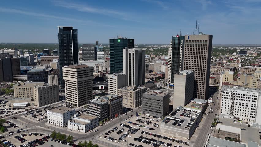 Downtown Winnipeg, Manitoba. High aerial drone moves toward the city skyline, revealing skyscrapers, streets, traffic, and the urban core under clear blue summer skies.