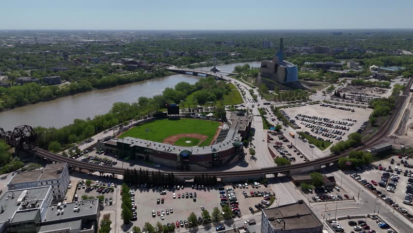 Winnipeg, Manitoba. Drone flies forward from Goldeyes stadium toward Red River and Canadian Museum for Human Rights with surrounding roads, buildings, and city skyline on a clear summer day.