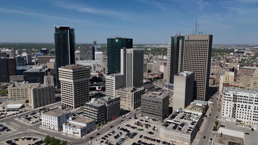 Downtown Winnipeg, Manitoba. Static drone shot overlooking Main Street, tall buildings, city streets, traffic, and distant horizon under clear blue summer skies.