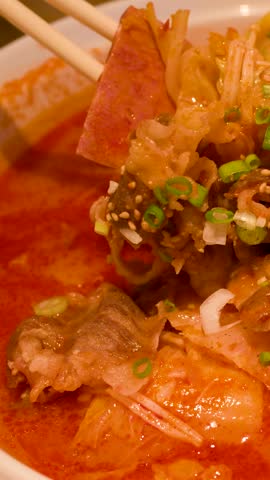 Close-up shot of chopsticks and spoon lifting beef and noodles from spicy red broth.