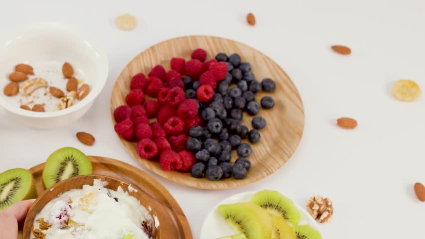 Overhead shot of hand scooping yogurt with berries, kiwi, and nuts from wooden bowl.