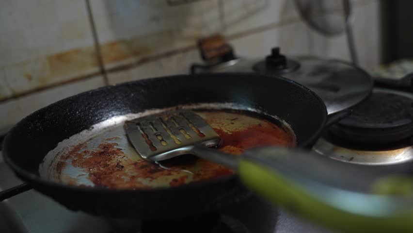 a dirty used frying pan with burnt grease and a metal spatula resting inside. It sits on a gas stovetop in a dimly lit domestic kitchen with a tiled background after cooking a meal.