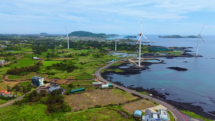 Aerial view of Sinchang windmill coastal road winding past wind turbines farmland and volcanic shoreline in Jeju Island South Korea