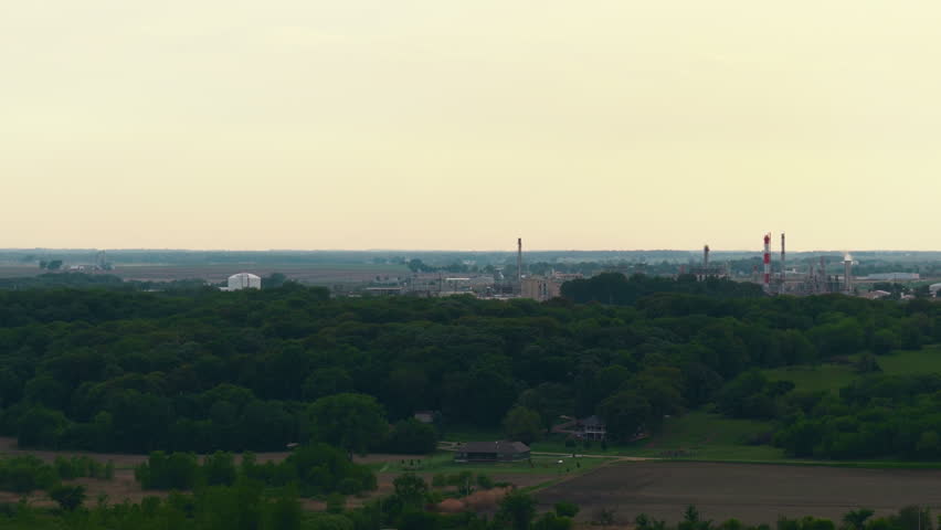 Woodland and farm fields toward a distant manufacturing complex with tall stacks on the horizon, under a vast pale sky in hazy summer light.
