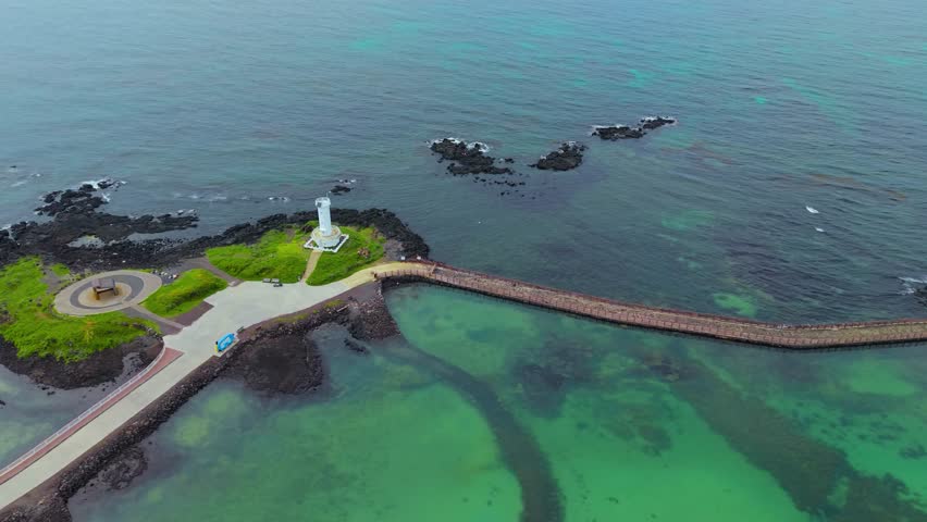 Aerial parallax of white Sinchang Lighthouse on rocky islet connected by walkway over emerald turquoise water Jeju Island Korea