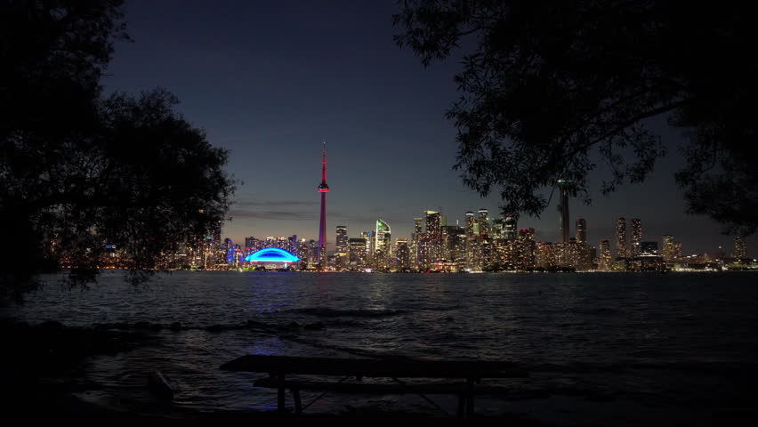 Toronto skyline and CN Tower illuminated at night as camera moves forward from trees toward the city waterfront, Slow Motion
