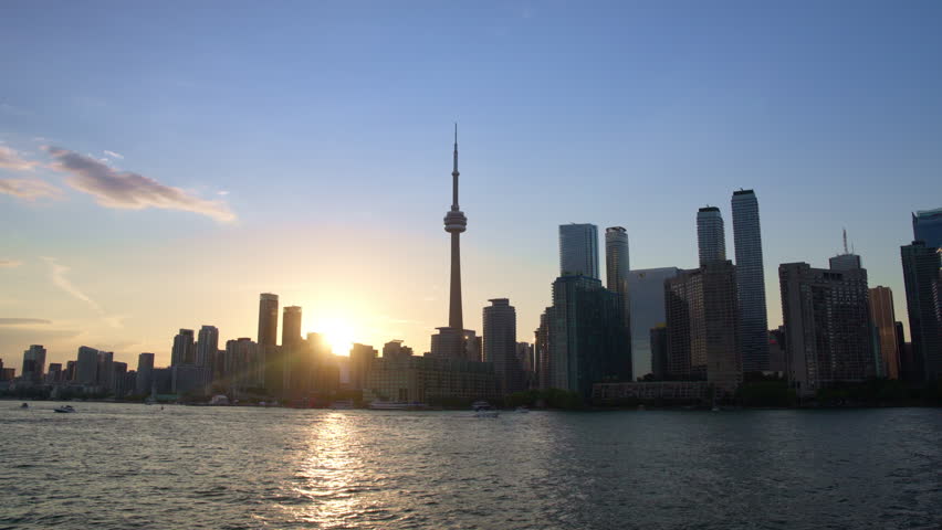 Wide view of Toronto skyline and CN Tower from a moving boat as the sun sets between buildings with reflections on the water, Slow Motion