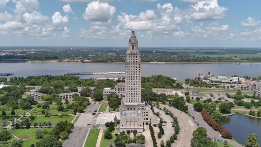 Dynamic aerial shot showcases Louisiana State Capitol building near Mississippi River. Features capitol building, river, green trees, and buildings under a cloudy sky.