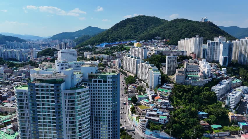 Aerial view of the urban landscape near Songdo Beach featuring high-rise residential buildings, dense city streets, and lush green hills in Busan, Busan, South Korea.