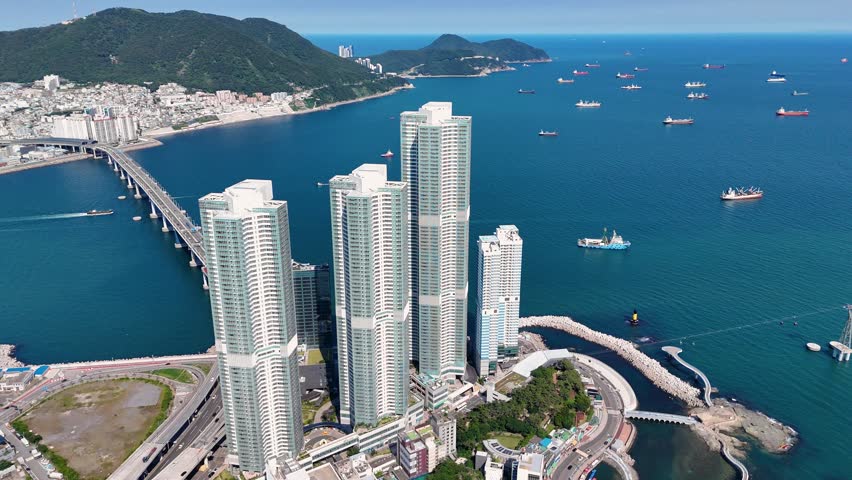 Aerial view of Songdo Beach featuring modern high-rise residential towers, the Namhang Bridge spanning the blue sea, and coastal roads in Busan, South Korea.