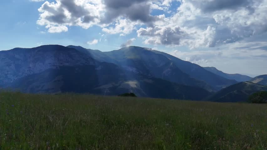 Mountain landscape with overlapping ridges illuminated by sunbeams filtering through the clouds, green meadow in the foreground and deep and bright natural atmosphere