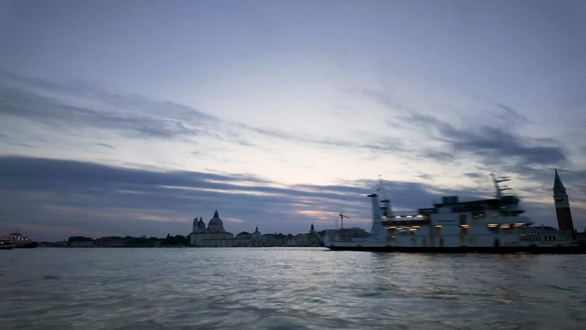 Day-to-night time-lapse of the Venice skyline across the Venetian lagoon. The sunset sky darkens as city lights illuminate historic landmarks including the Basilica della Salute and St. Mark